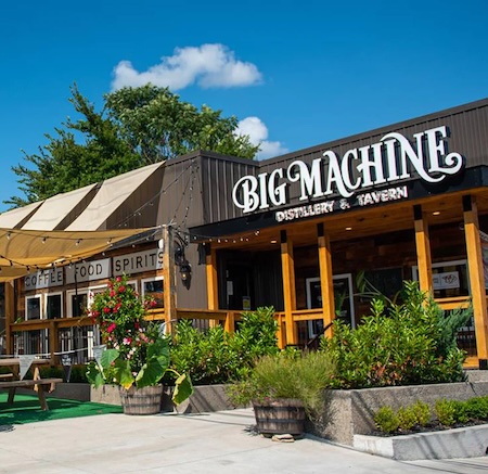 Exterior view of Big Machine Distillery & Tavern with wooden structure, outdoor seating, and vibrant greenery under a clear blue sky.
