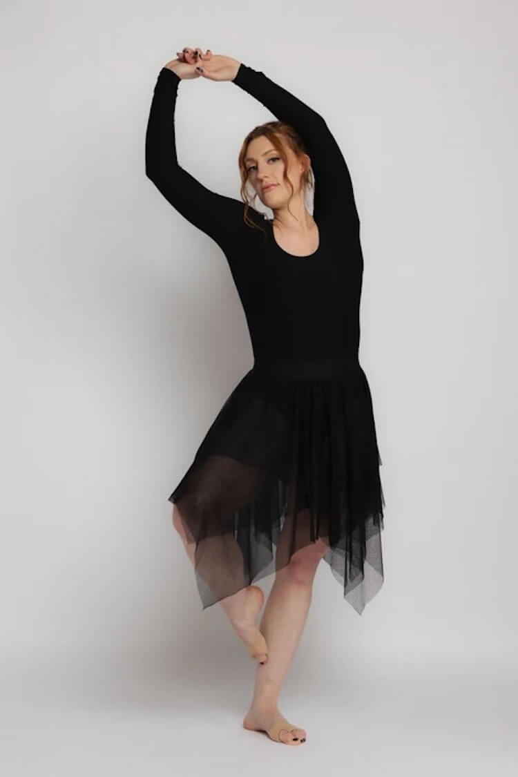 Woman in black long-sleeve leotard and sheer black skirt posing with arms raised and one foot crossed barefoot.